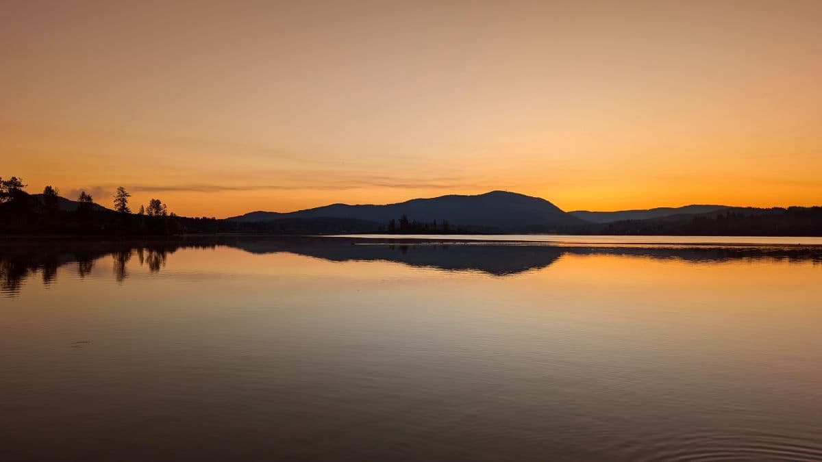 Photo of a Lake in Cowichan Valley at sunrise on a clear morning. The mountains and trees reflect off the calm water of the lake producing an image that looks like a sound wave form.