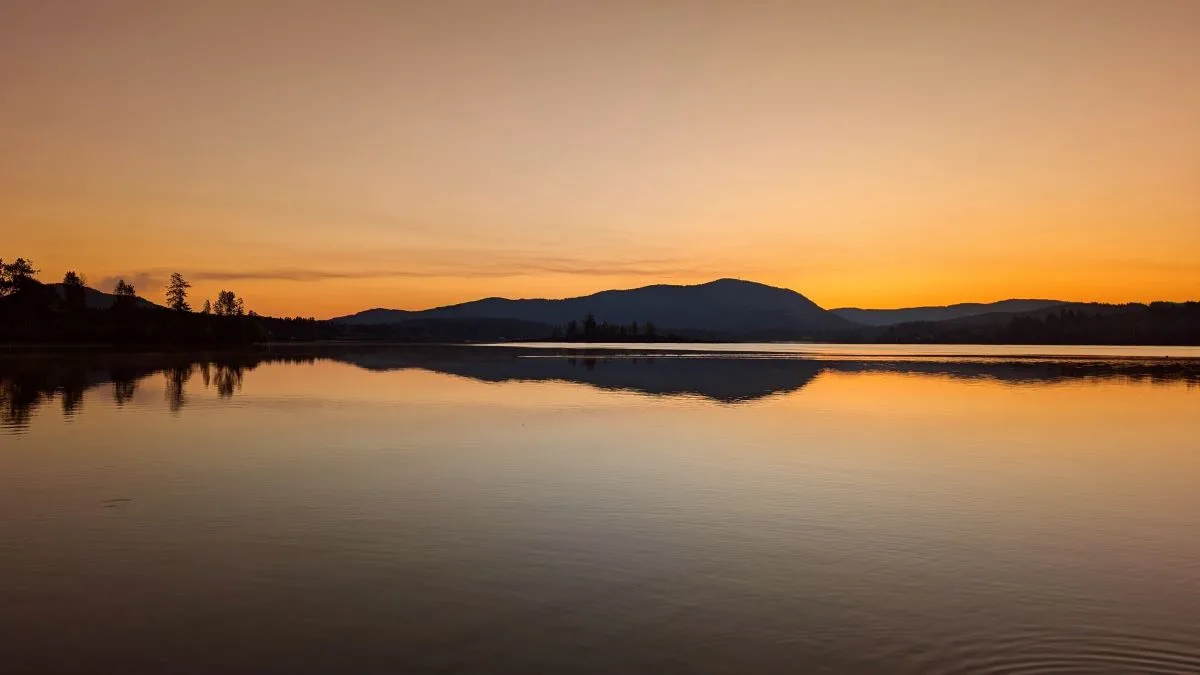 Photo of a Lake in Cowichan Valley at sunrise on a clear morning. The mountains and trees reflect off the calm water of the lake producing an image that looks like a sound wave form.